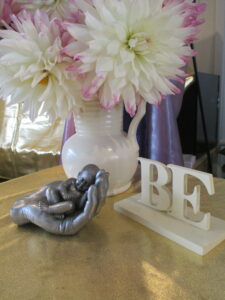 A tabletop arrangement featuring a sculpted ornament spelling “BE,” a white jug filled with fresh flowers, and a silver cast of hands cradling a baby.